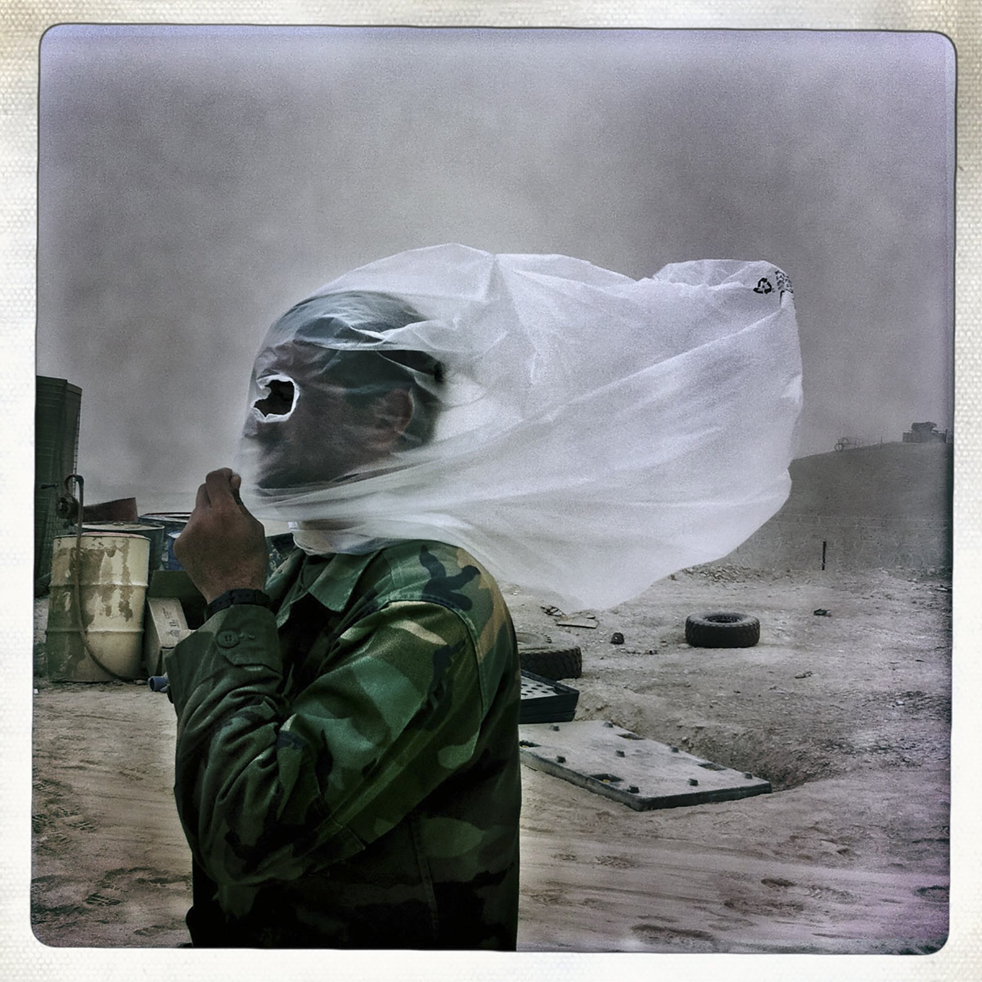 L'image montre un soldat portant un uniforme camouflé, protégé par un morceau de tissu fin qui couvre son visage. Le vent soulève le tissu, créant une atmosphère dramatique. En arrière-plan, le paysage semble poussiéreux et désertique, avec des objets flous tels que des pneus. L'ensemble de la scène évoque un contexte difficile, probablement lié à des conditions climatiques extrêmes.