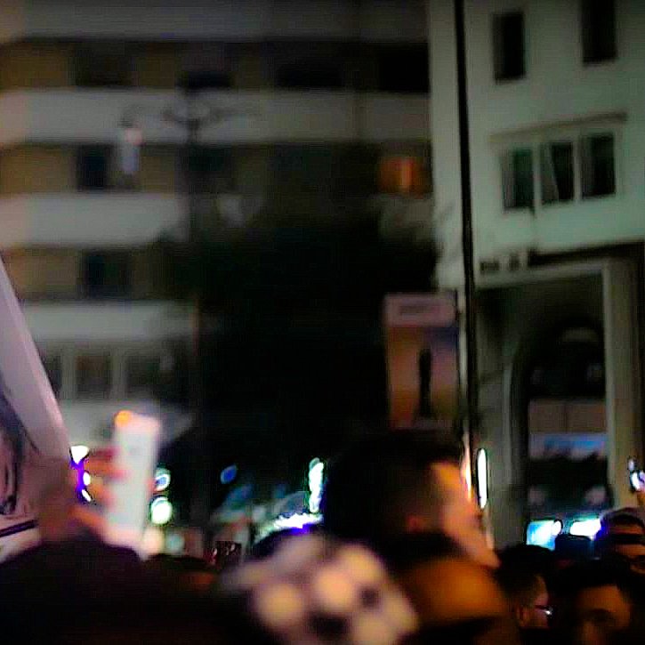 تظهر الصورة مجموعة من الأشخاص في مظاهرة أو تجمع حاشد ليلي. في المقدمة، يرفع أحد المشاركين لوحة تحمل صورة شخص، بينما يظهر جزء من الحشود في الخلفية. يبدو أن الأضواء والمباني حولهم تخلق جوًا حضريًا يعكس الحركة والنشاط. تعبر الصورة عن تعبير قوي عن الآراء والمشاعر الجماعية.