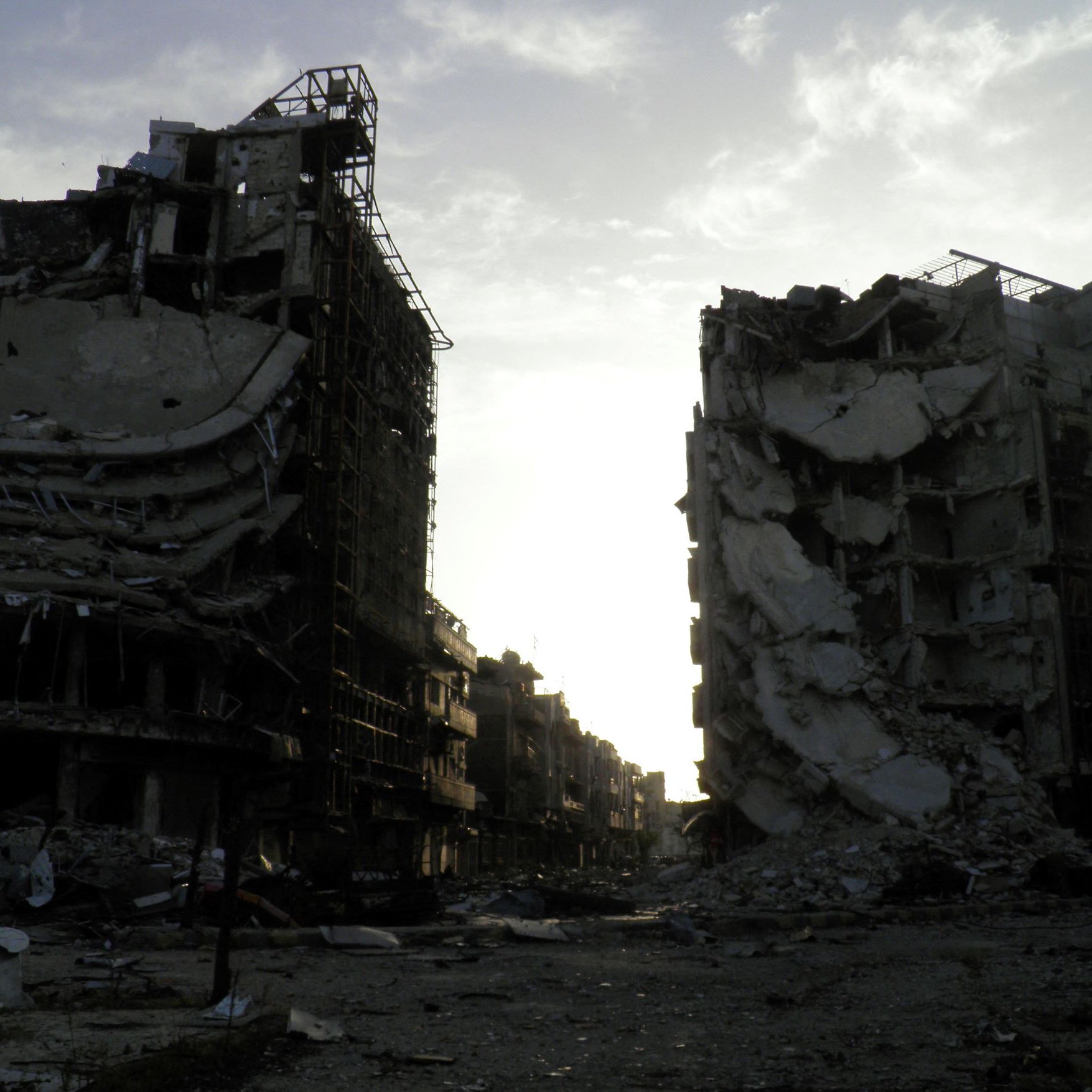 L'image montre deux bâtiments en ruines, témoignant des conséquences d'un conflit ou d'une catastrophe. Les structures sont partiellement effondrées, avec des débris dispersés autour. L'éclairage est sombre, suggérant un crépuscule ou une journée nuageuse, ce qui ajoute une atmosphère de désolation à la scène. Le paysage environnant semble aussi dévasté, accentuant la gravité de la situation.