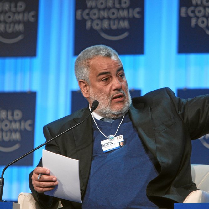 L'image montre un homme en train de parler lors d'un événement, probablement une conférence ou un forum. Il porte un costume noir avec un pull bleu en dessous. Il semble être engagé dans une discussion, levant le bras pour souligner un point. En arrière-plan, on peut voir des panneaux portant le logo du Forum économique mondial. L'ambiance paraît professionnelle et dynamique.