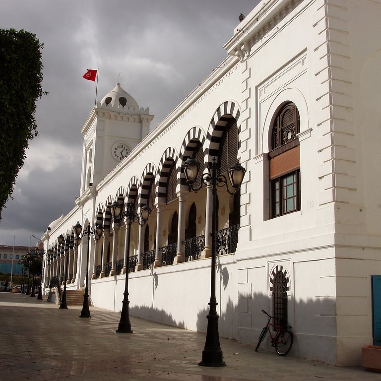 L'image montre un bâtiment architectural imposant, probablement un hôtel de ville ou un bâtiment administratif, avec une façade blanche. Ce bâtiment présente des arches décoratives et des fenêtres ornées. On peut apercevoir une grande horloge sur la tour, ainsi qu'un drapeau turc flottant au sommet. Le ciel est nuageux, ajoutant une ambiance dramatique à la scène. Des lampadaires sont alignés le long de la rue pavée qui borde le bâtiment, et un vélo est garé sur le côté. Des arbres bien taillés sont également visibles, contribuant à l'esthétique générale de l'endroit.