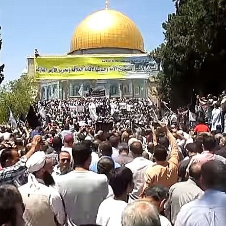L'image montre une foule rassemblée devant un bâtiment orné d'un dôme doré. Les participants semblent être engagés dans une manifestation ou un rassemblement, avec des drapeaux et des pancartes visibles. Les arbres environnants ajoutent au décor, et l'atmosphère générale paraît dynamique, marquée par une forte présence de personnes.