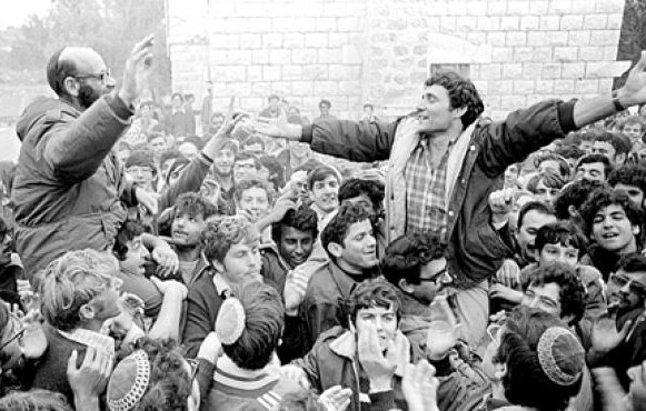 L'image montre une foule de personnes rassemblées, avec des visages enthousiastes et des expressions de joie. Deux hommes se distinguent, l'un portant des lunettes, debout sur les épaules de l'autre, qui lève les bras en signe de célébration. Les membres de la foule semblent excités et engagés, et l'environnement est peut-être urbain ou rural, entouré de bâtiments en pierre. L'atmosphère générale évoque un moment de rassemblement collectif et de camaraderie.