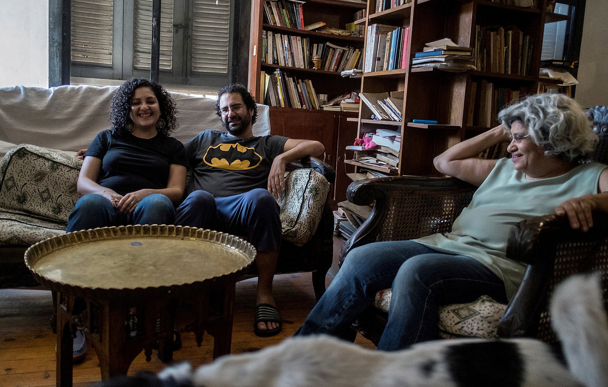 L'image montre un groupe de trois personnes assises dans un salon. Il y a deux femmes et un homme. La première femme, à gauche, sourit et porte un t-shirt noir. L'homme, au centre, porte un t-shirt avec le logo de Batman. La deuxième femme, à droite, a les cheveux bouclés et semble détendue. Ils sont assis sur des canapés, entourés d'étagères remplies de livres et d'objets décoratifs. Une ambiance conviviale et décontractée se dégage de la scène.