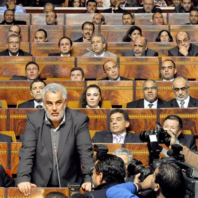 L'image montre une séance dans un bâtiment législatif, où une personne (probablement un membre du gouvernement ou un habile orateur) se tient devant un grand groupe de personnes assises. Les membres de l'assemblée, en arrière-plan, semblent concentrés sur le discours prononcé. On peut voir des journalistes avec des caméras, suggérant que cet événement est couvert par les médias. L'atmosphère est formelle et sérieuse, typique de débats ou de discussions politiques.