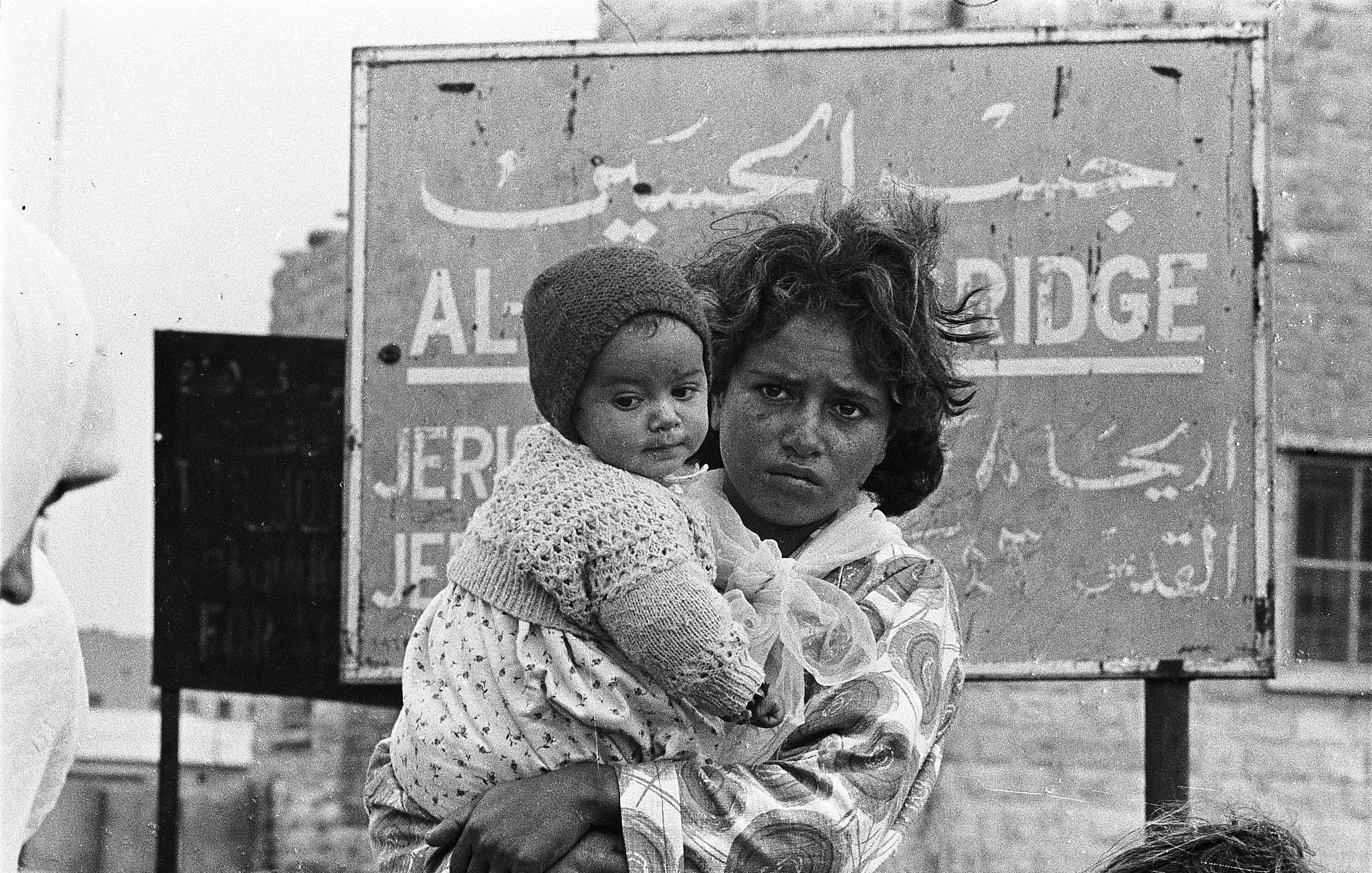 این تصویر یک کودک را نشان می‌دهد که در آغوش یک زن ایستاده است. زن ظاهراً نگران و جدی به نظر می‌رسد و کودک در آغوشش پایش را به دور او حلقه کرده است. پس‌زمینه تصویر شامل یک تابلو با نوشته‌ها به زبان عربی و انگلیسی است که نام مکانی را نشان می‌دهد. حالت چهره و لباس‌های آن‌ها نشان‌دهنده سختی‌های احتمالی در زندگی است. تصویر به وضوح وضعیت انسانی و احساسات عمیق را منتقل می‌کند.