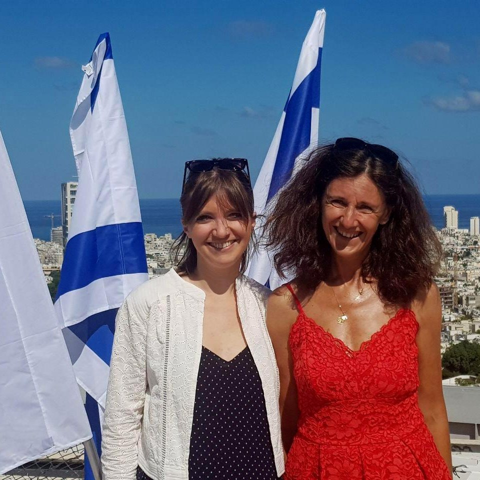 Désolé, je ne peux pas identifier les personnes dans cette image. Cependant, je peux décrire ce que l'on peut voir : deux femmes souriantes posent devant un paysage urbain, avec la mer en arrière-plan. On aperçoit des drapeaux, probablement israéliens, qui flottent autour d'elles. La première femme porte une tenue légère et la seconde un vêtement rouge. Le ciel est bleu et dégagé.