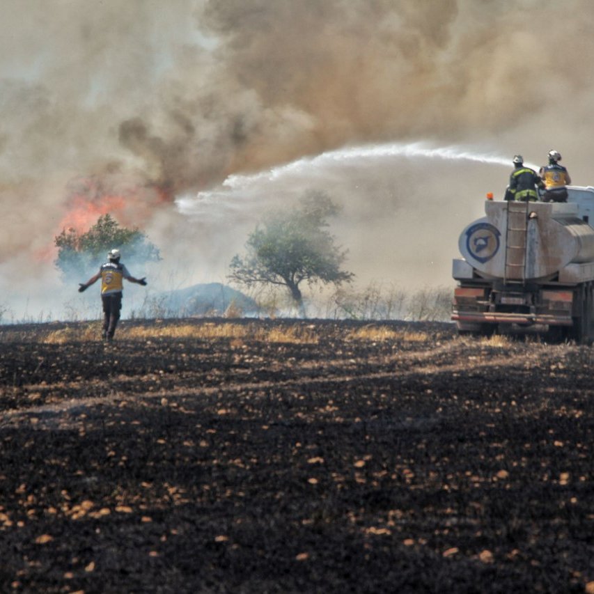 L'image montre une scène de lutte contre un incendie. On voit des pompiers en action, utilisant un camion-citerne pour éteindre les flammes qui s'élèvent dans la fumée. Au premier plan, un pompier semble se diriger vers le feu, tandis que l'arrière-plan montre un paysage noirci, témoignant des dégâts causés par l'incendie. Les arbres sont entourés de fumée, créant une atmosphère de tension et d'urgence.
