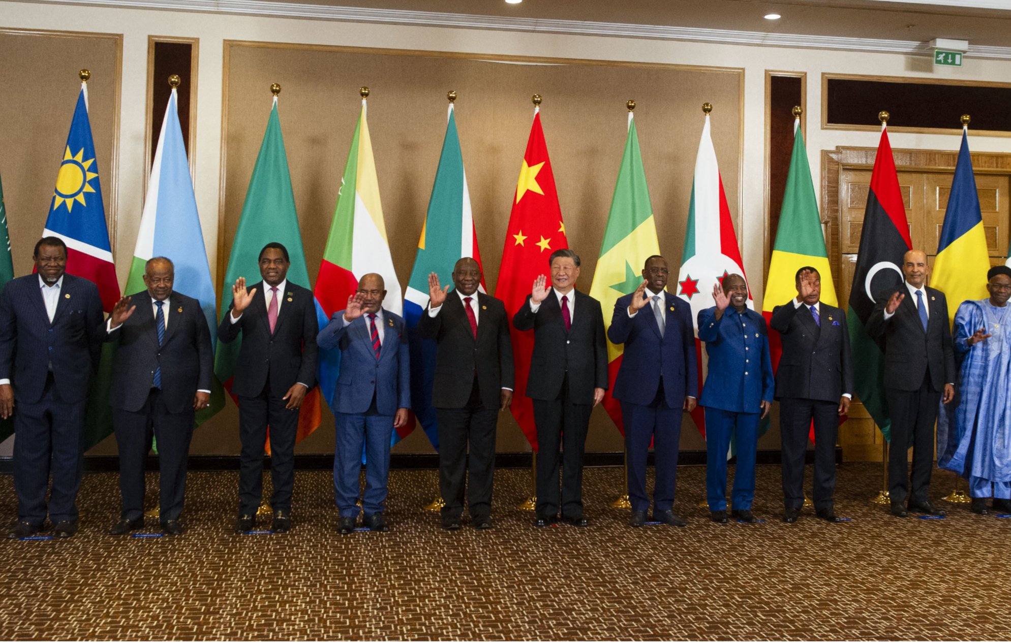 La imagen muestra a un grupo de líderes y representantes de diferentes países alineados en una sala. Detrás de ellos hay banderas de varias naciones, incluidos países africanos y de otras partes del mundo. Todos los individuos parecen estar posando para una fotografía, algunos levantando la mano en señal de saludo. La decoración del lugar es elegante, con un ambiente formal que refleja la importancia del evento.