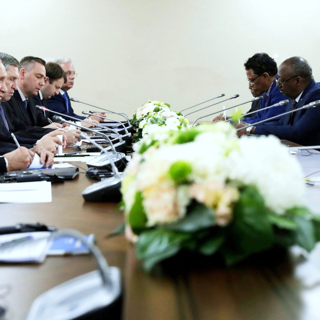 L'image représente une réunion ou une conférence avec plusieurs participants assis autour d'une grande table. Au centre de la table, il y a un arrangement floral, qui ajoute une touche de décoration. Les participants semblent être concentrés et engagés dans la discussion, prenant des notes ou écoutant attentivement. L'atmosphère est sérieuse et professionnelle, suggérant un dialogue important entre les différentes parties présentes.
