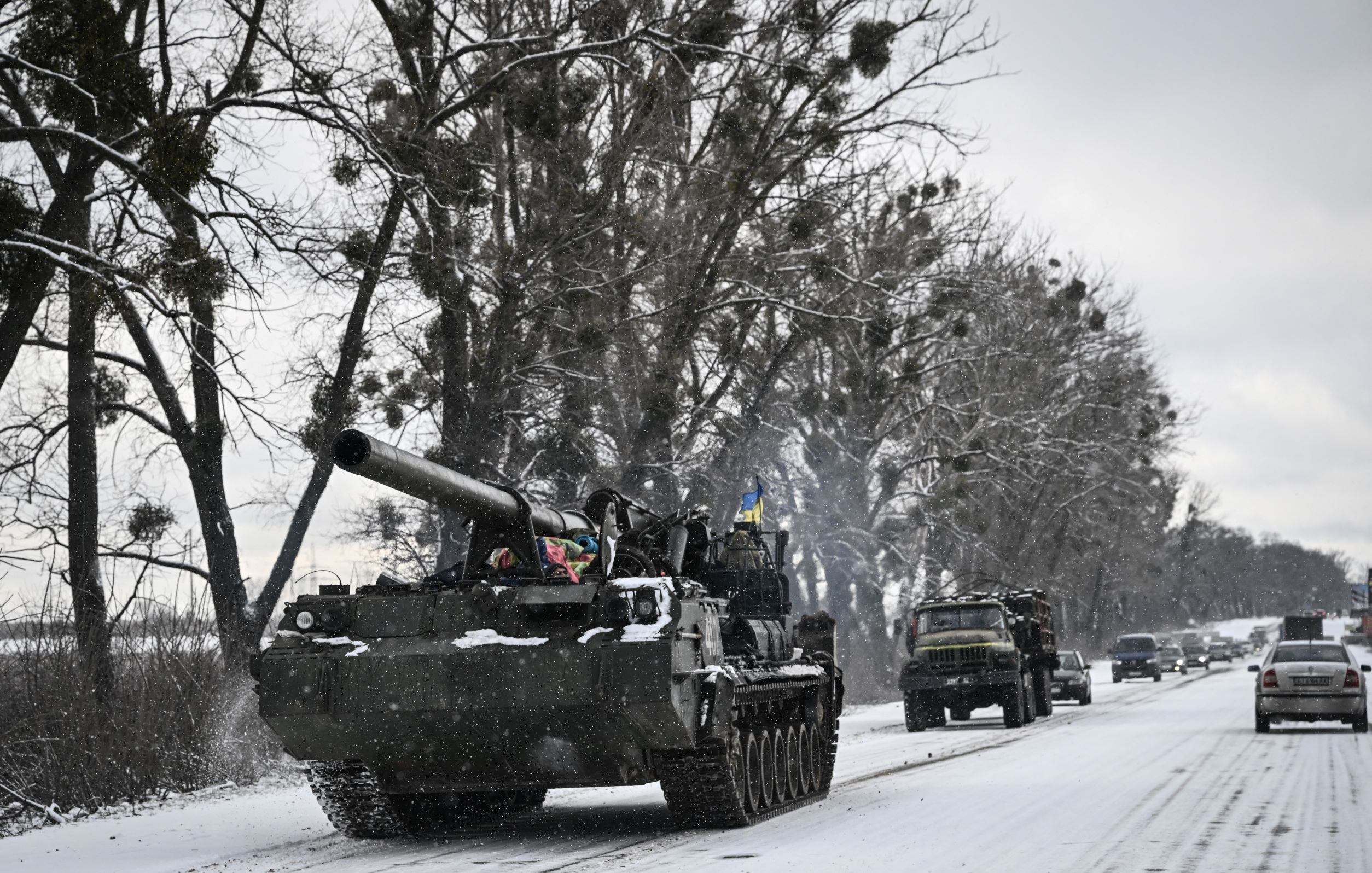 L'immagine mostra un carro armato che si muove lungo una strada innevata, circondato da alberi. Il carro armato ha una canna protrudente e presenta dettagli decorativi che potrebbero indicare l'appartenenza a una specifica unità militare. Accanto al carro armato, si intravedono anche alcuni veicoli, probabilmente militari, che viaggiano sulla stessa strada. Il paesaggio è caratterizzato da un'atmosfera invernale, con la neve che copre sia la strada che il terreno circostante.