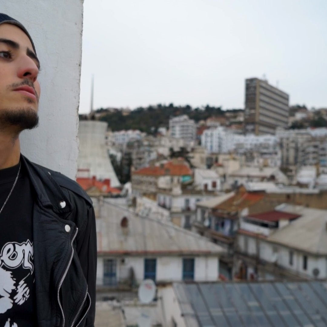 L'image montre un jeune homme qui se tient sur un toit, regardant au loin. Il porte un bonnet et une veste en cuir noire, ainsi qu'un t-shirt avec un design graphique. En arrière-plan, on aperçoit une ville avec des bâtiments de différentes tailles et architectures, et des toits de maisons. Le ciel est légèrement nuageux, ce qui donne une ambiance calme et contemplative à la scène.