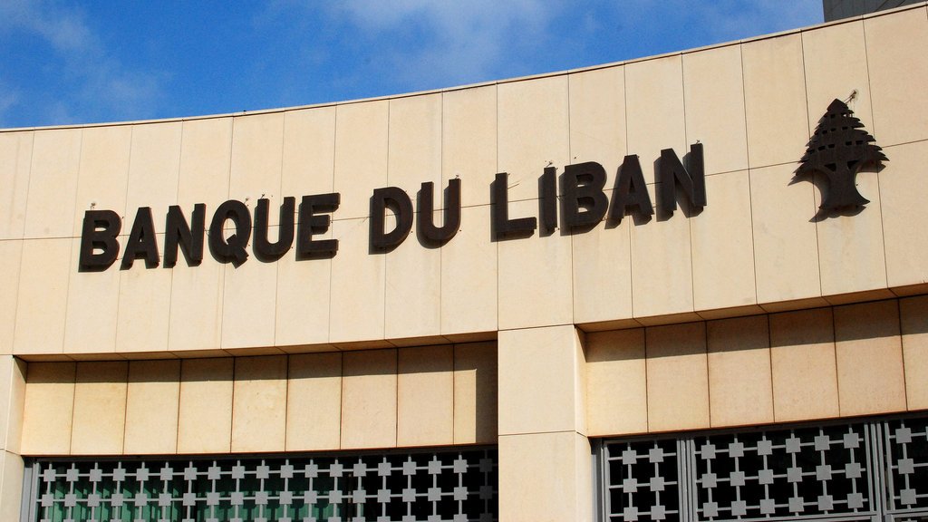 تظهر الصورة واجهة مؤسسة مالية تحمل اسم "بنك لبنان" بالخط العريض. الكتابة باللون الأسود على خلفية فاتحة، وتوجد شجرة الأرز، وهي رمز وطني للبنان، بجانب الاسم. السماء زرقاء مع بعض السحب، مما يعطي انطباعًا بوجود يوم مشمس. يبدو أن الجزئية المعروضة من المبنى حديثة ومرتبة.