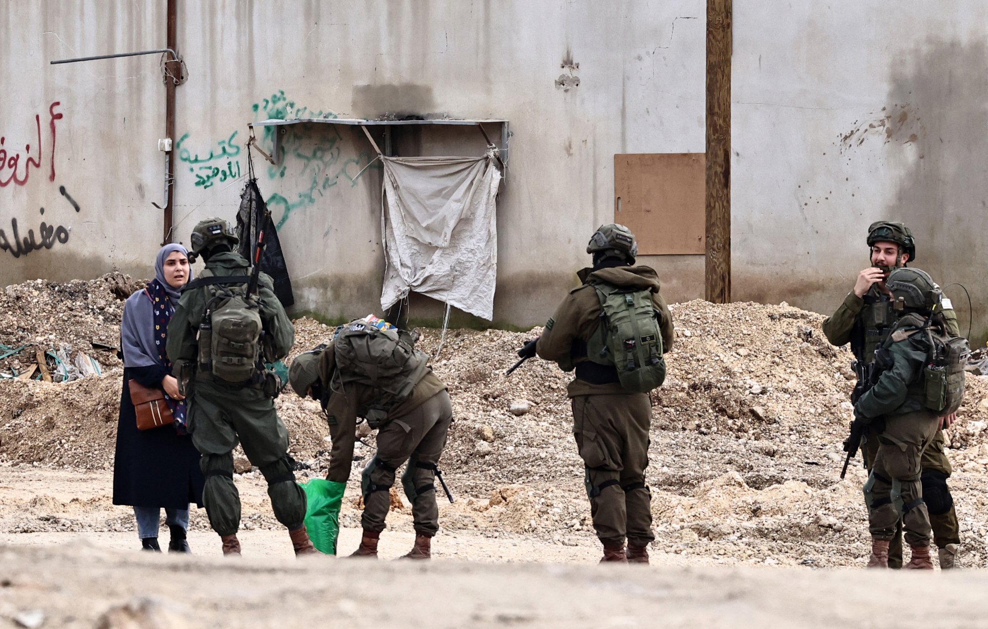 L'image montre une femme vêtue d'une robe sombre, se tenant à côté d'une rue. Devant elle, plusieurs soldats en uniforme sont regroupés, certains sont en train de fouiller un sac, tandis que d'autres observent la femme. En arrière-plan, on peut voir un mur avec des graffitis et des débris sur le sol.