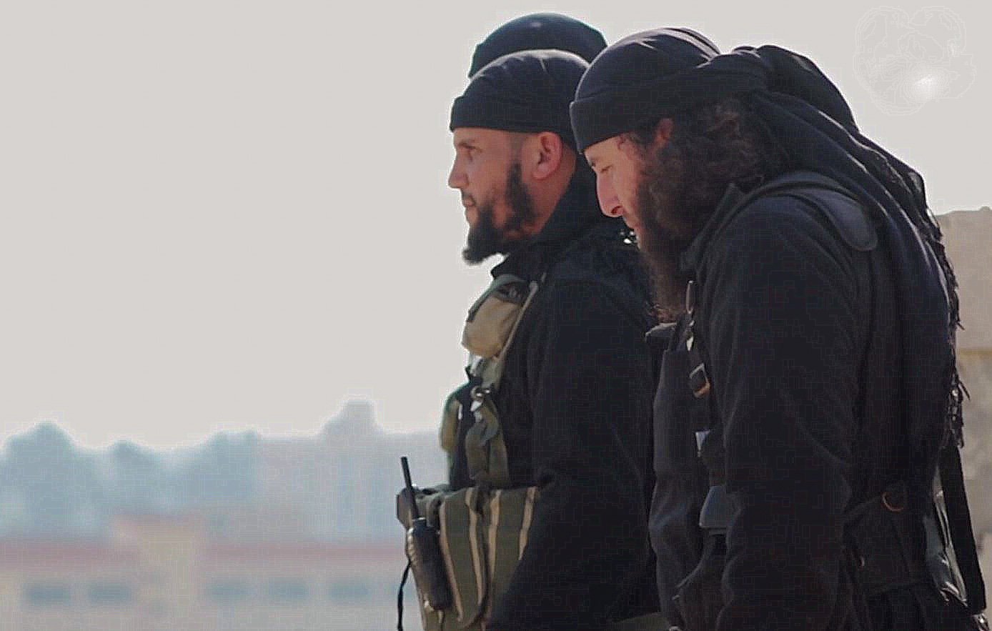 L'image montre deux hommes en tenue militaire, portant des casques et des écharpes noires. Ils ont des barbes et semblent être en train de surveiller les environs. L'arrière-plan est flou, ce qui donne une impression de concentration sur les personnages au premier plan. L'atmosphère semble sérieuse et tendue.