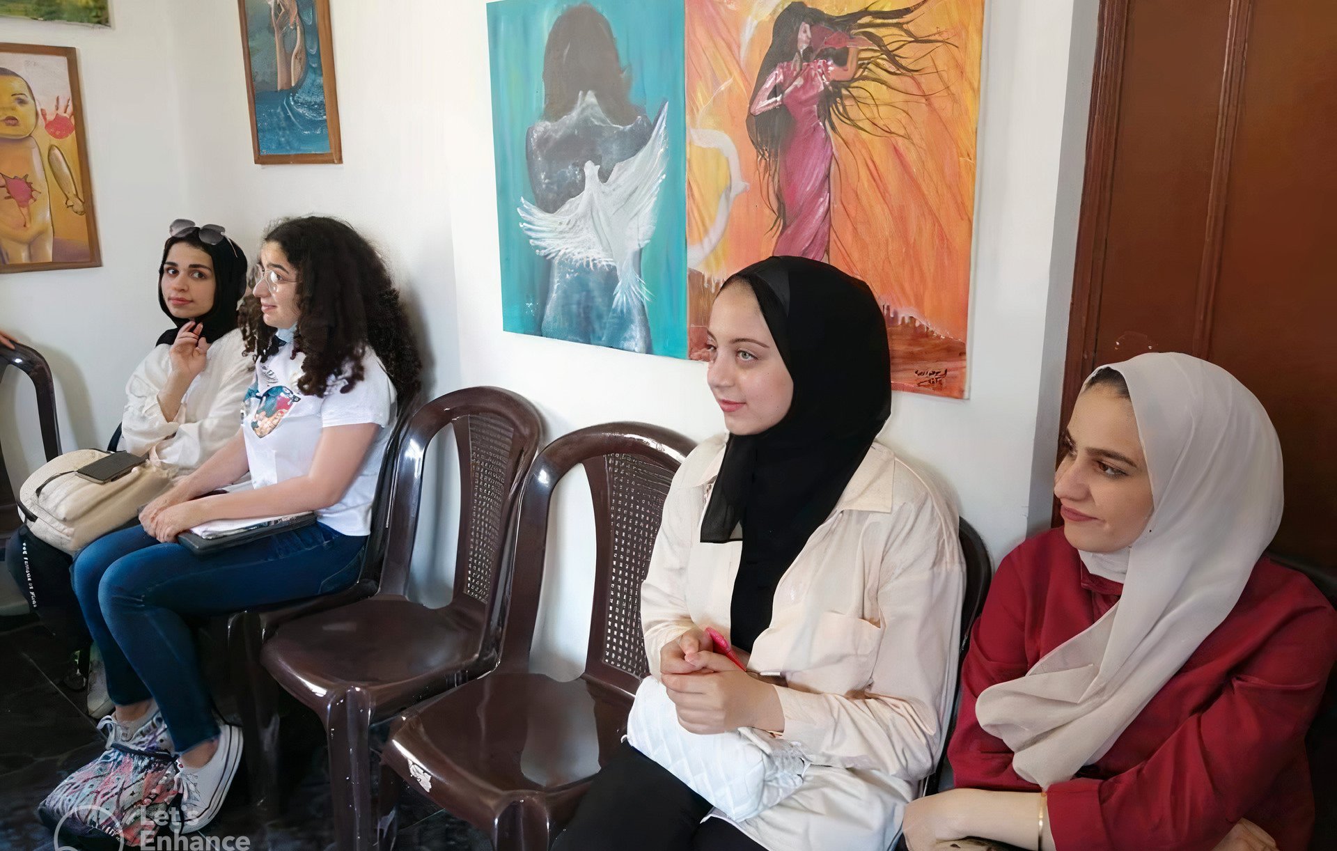 Nell'immagine si vedono quattro donne sedute su delle sedie in una stanza. Le pareti sono decorate con opere d'arte colorate, rappresentanti differenti stili. Le donne indossano abbigliamenti diversi: alcune hanno il velo, altre hanno abiti casual. Il loro atteggiamento sembra tranquillo, mentre osservano l'ambiente circostante. La luce che entra dalla finestra illumina la scena, creando un'atmosfera accogliente.