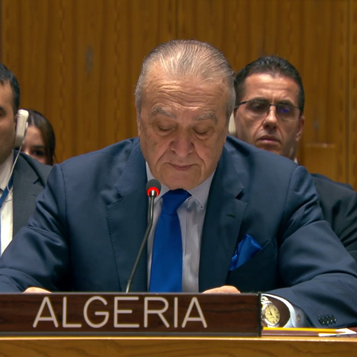 Un homme en costume parle à un podium, des personnes écoutent derrière lui.