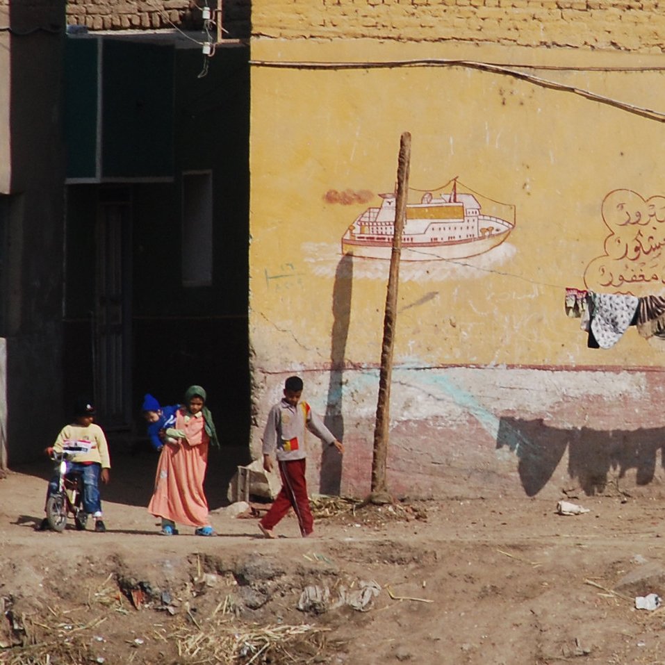 L'image montre une scène de rue dans un environnement rural ou semi-urbain. On peut y voir plusieurs personnes, dont des enfants, qui se déplacent. À l'arrière-plan, il y a des bâtiments avec des murs peints, incluant des motifs ou des inscriptions. Des vêtements sont suspendus à une corde, presque comme un étendage. Le sol est poussiéreux, et l'ensemble de la scène dégage une atmosphère de vie quotidienne dans un cadre modeste.