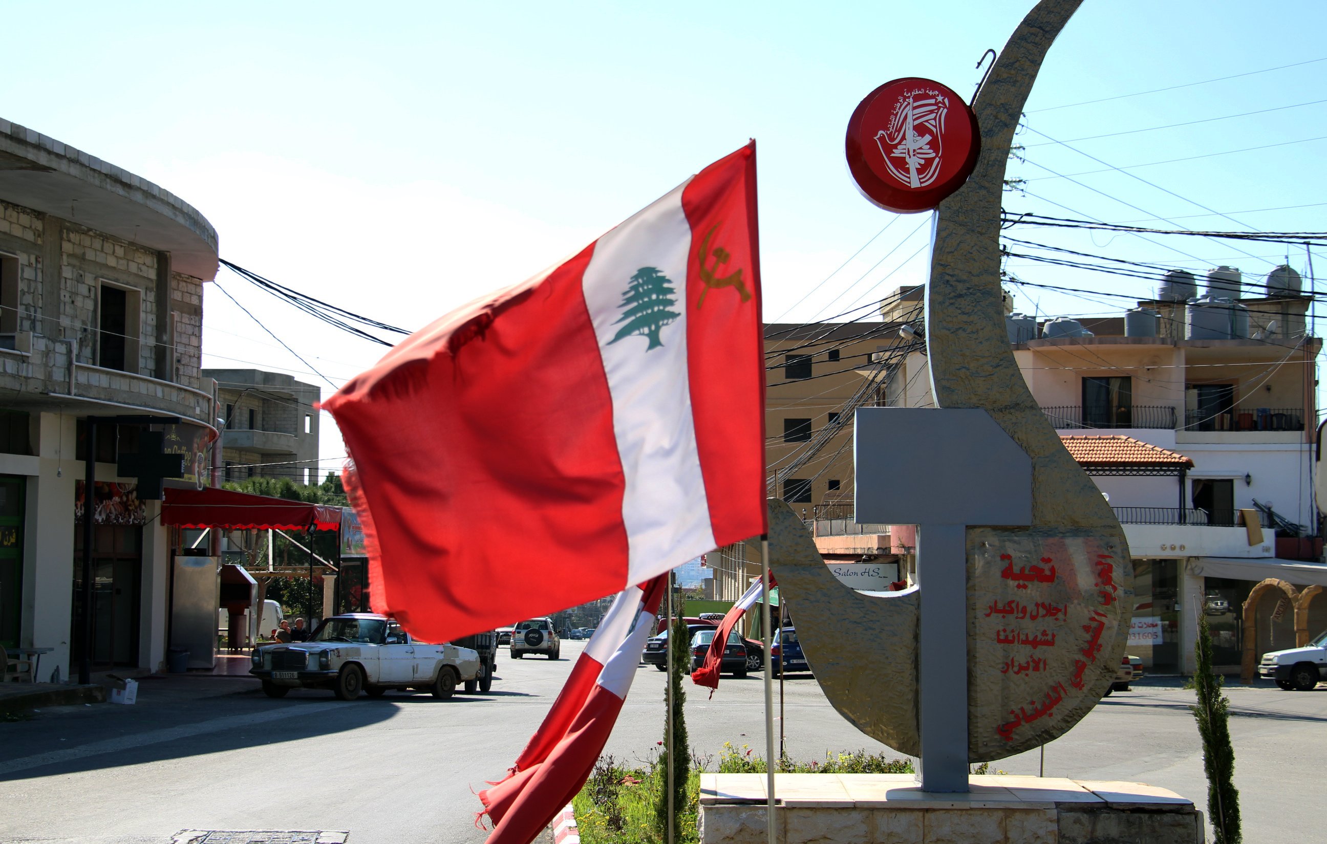 La imagen muestra una intersección en una localidad, con una bandera de Líbano ondeando en el viento. A la derecha, hay una estructura que parece una escultura o un monumento, posiblemente con inscripciones en árabe. Al fondo se pueden ver edificios y postes de luz, mientras que el cielo es despejado y soleado. La escena refleja un ambiente urbano característico de la región.