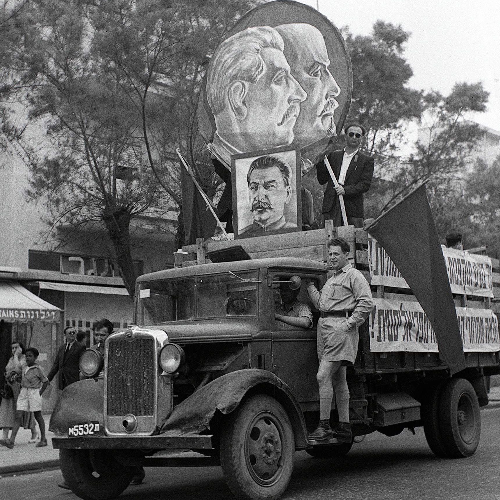 L'image représente un défilé historique avec un camion décoré. Sur le camion, on peut voir des portraits de figures politiques, probablement des leaders communistes, entourés de drapeaux. Un homme en uniforme se tient debout sur le camion, tandis que d'autres personnes défilent en arrière-plan. L'atmosphère semble festive et engagée, typique des manifestations politiques d'époque. Les bâtiments et les passants en arrière-plan ajoutent un contexte urbain à la scène.