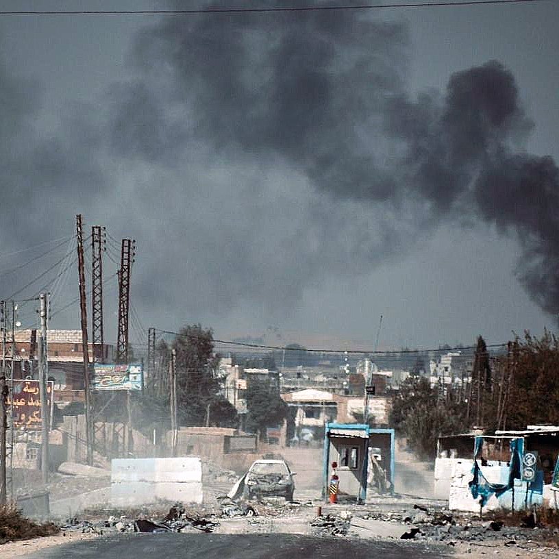 The image depicts a desolate street under a dark, smoke-filled sky. In the background, billowing plumes of black smoke suggest an ongoing fire or explosion. The foreground shows a damaged road with debris, potentially hinting at a conflict zone. There are signs of destruction, including burnt vehicles and abandoned structures, contributing to an overall atmosphere of turmoil and devastation. The landscape appears barren with a few power lines and distant buildings, reflecting the impact of violence on the area.