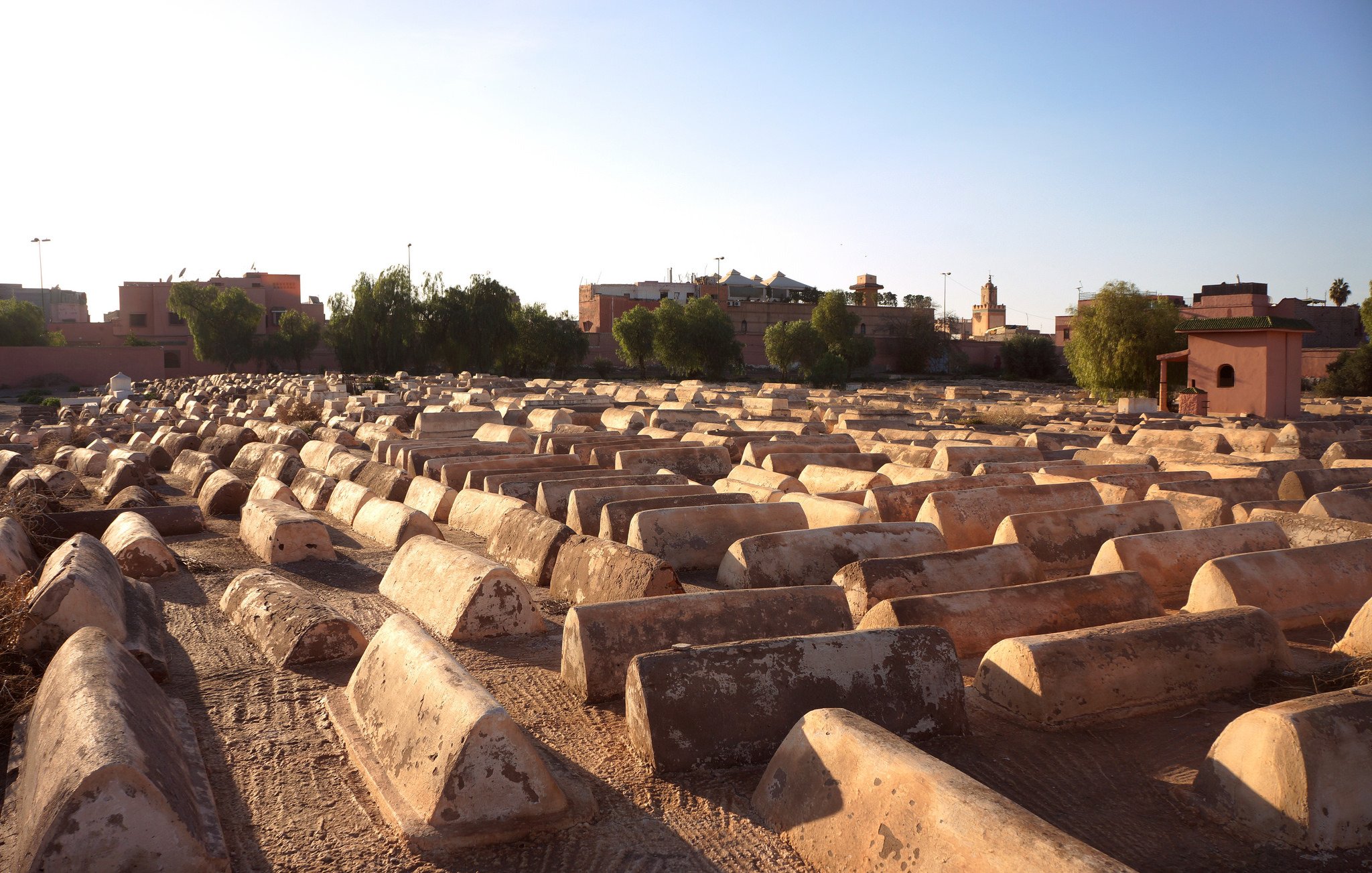 تظهر الصورة منظرًا لمقبرة كبيرة، حيث تتوزع القبور بشكل منظم على الأرض. تبدو القبور مصنوعة من الحجر أو الطين، وبعضها يظهر عليه آثار الزمن. في الخلفية، هناك مباني ومدن صغيرة تظهر تحت ضوء الشمس، مما يعطي إحساسًا بالهدوء والسكينة. السماء تبدو صافية، مما يضيف إلى جمالية المشهد.