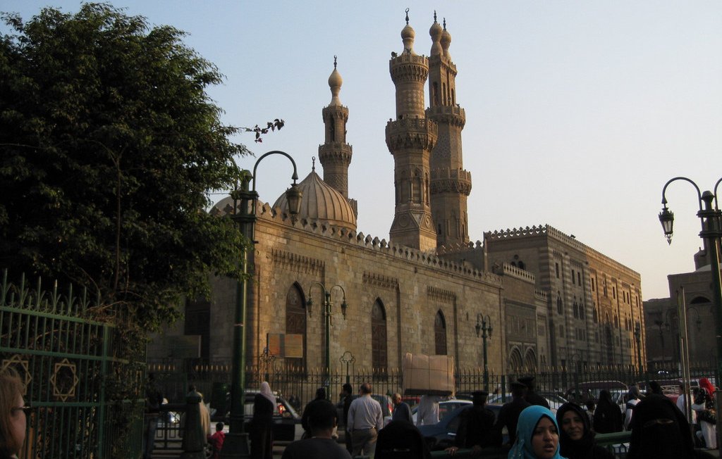 L'image montre un vaste bâtiment architectural au style islamique, probablement une mosquée, avec des minarets majestueux s'élevant vers le ciel. On peut voir des personnes en tenue traditionnelle, certaines portant le hijab. En arrière-plan, la lumière du soleil est douce, créant une atmosphère paisible. Les détails architecturaux de la façade en pierre ajoutent une touche historique à la scène. Des arbres et des lampadaires embellissent également l'environnement.
