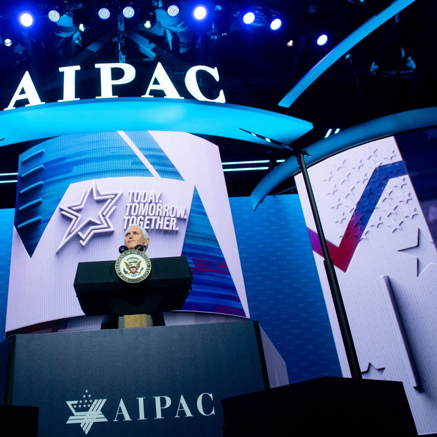 تظهر في الصورة منصة تتزين بشعار "AIPAC" (اللجنة الأمريكية للعلاقات العامة مع إسرائيل) مع إضاءة زرقاء مميزة. خلف المنصة، يوجد شعار يحمل عبارة "Today. Tomorrow. Together."، مما يشير إلى فعالية أو مؤتمر قادم. كما تتواجد شعارات وديكورات مضاءة أخرى تساهم في خلق جو حيوي ونشيط.