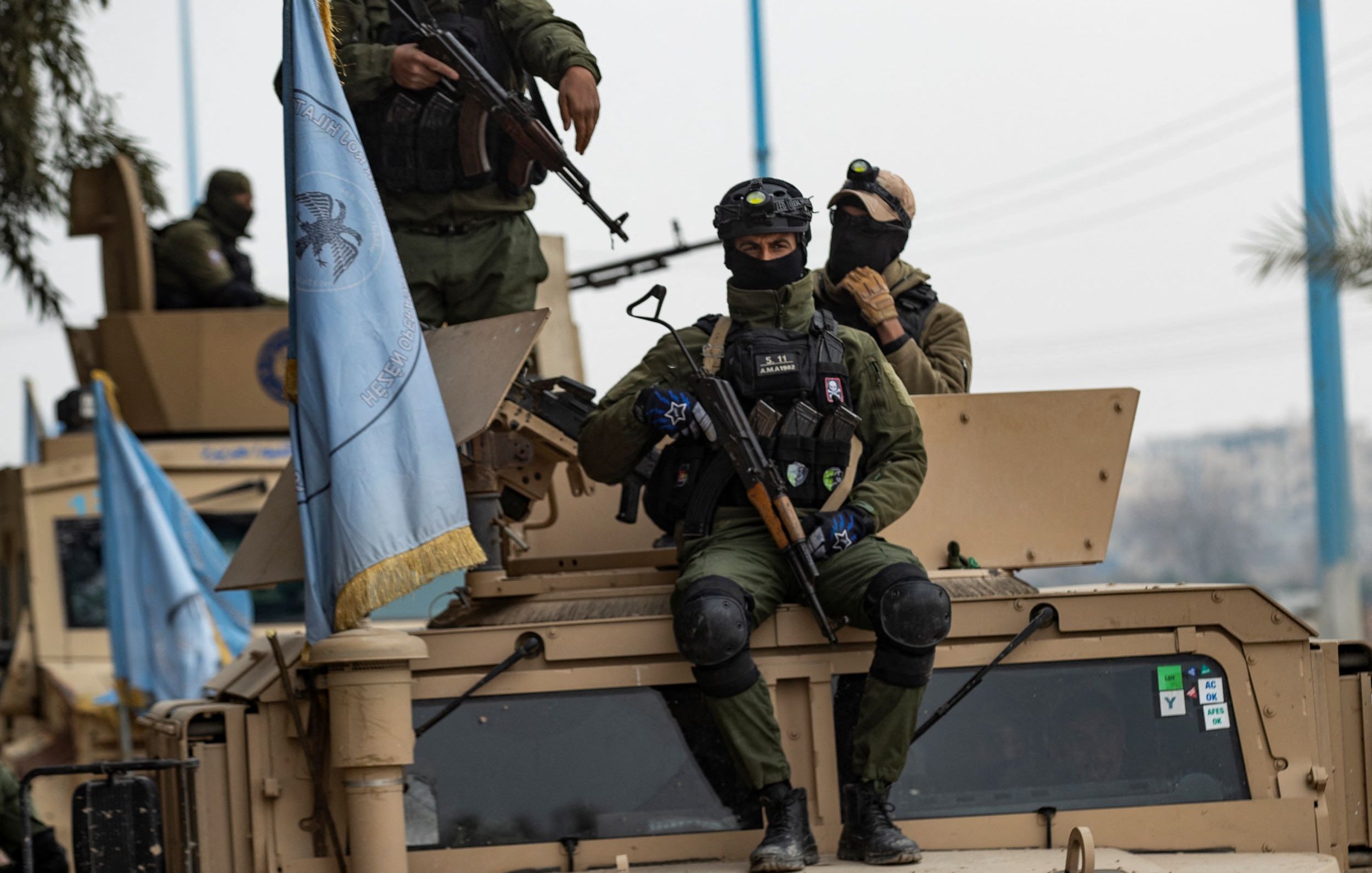 L'image montre des soldats en uniforme, armés et équipés de casques, se trouvant sur un véhicule militaire. L'un des soldats est assis sur le toit du véhicule, tenant une arme, tandis que d'autres soldats sont debout à proximité. On peut également voir un drapeau, probablement lié à une mission de maintien de la paix. L'environnement semble urbain, avec des éléments de la ville en arrière-plan.