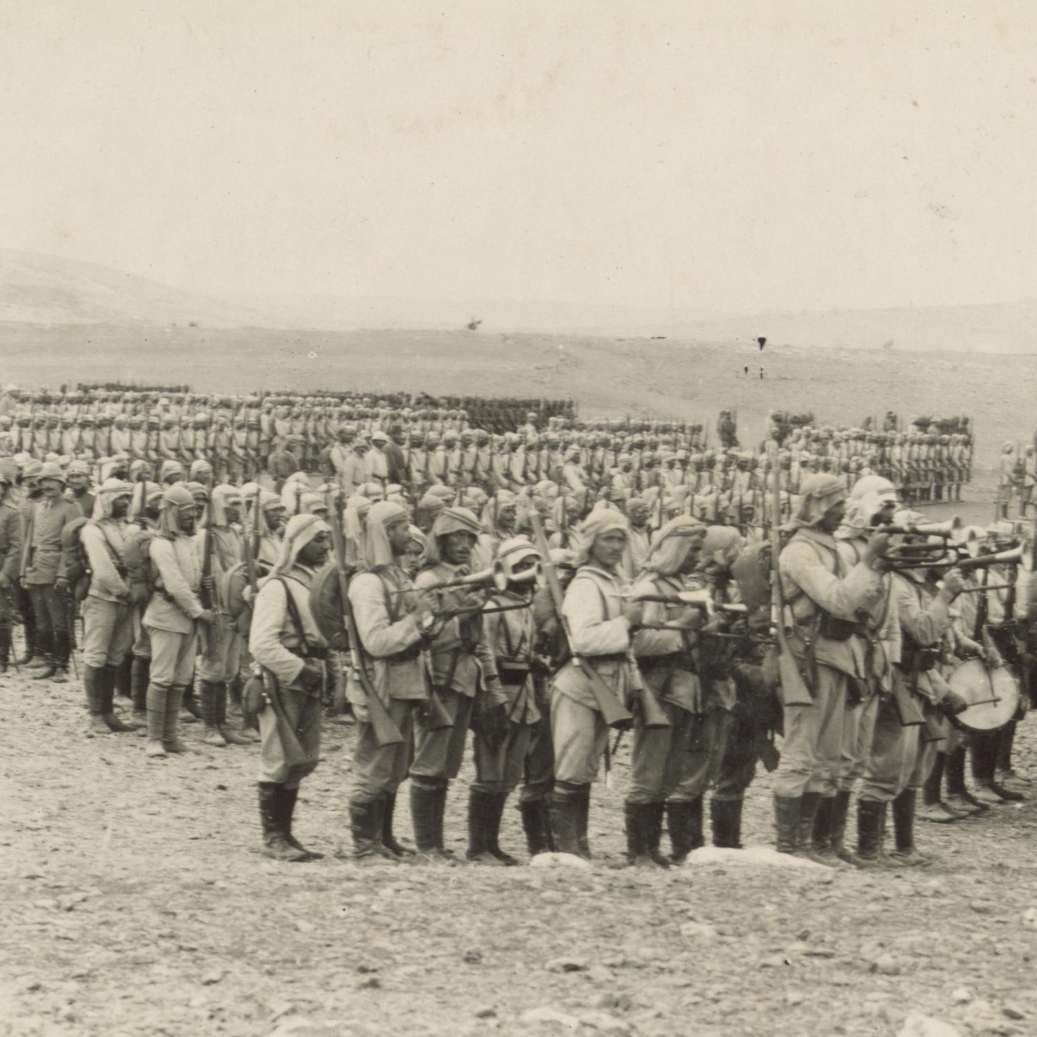 L'image montre une grande formation de soldats en uniforme, probablement d'un conflit militaire historique. Ils sont alignés en rangées serrées sur un terrain désertique ou peu végétalisé. Certains soldats portent des armes et semblent être en position de marche ou de formation. En arrière-plan, on peut voir d'autres soldats formant également des rangées, ce qui donne une impression de mobilisation de grandes troupes. L'atmosphère est sérieuse et solennelle, évoquant le contexte militaire de l'époque.