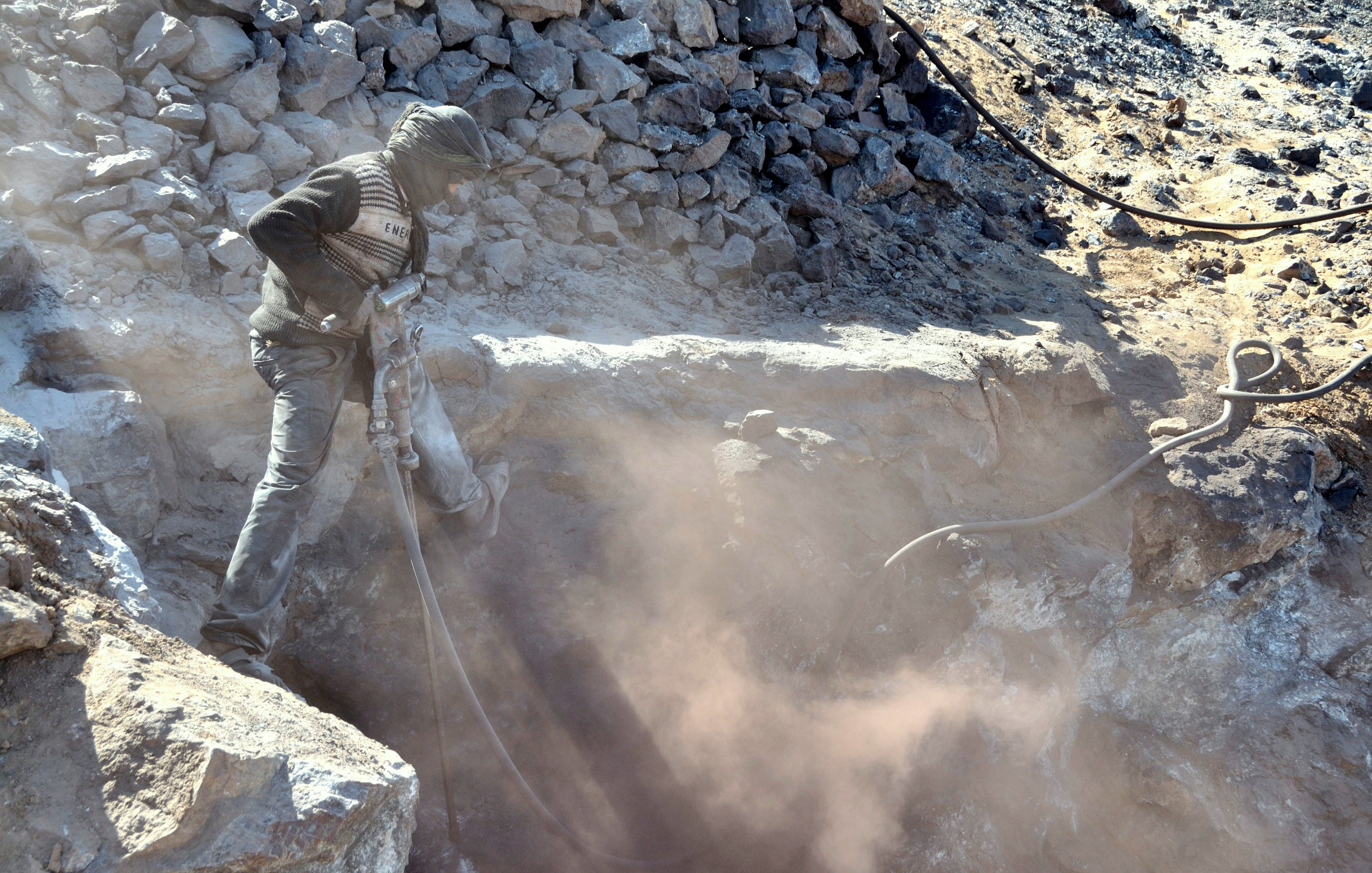 The image depicts a worker operating a pneumatic drill in a rocky area. The person is wearing protective clothing, including a mask or scarf over their face to shield against dust. Surrounding the worker are loose rocks and debris, and a cloud of dust is being kicked up by the drilling activity, creating a hazy atmosphere. The setting appears to be a mining or construction site.