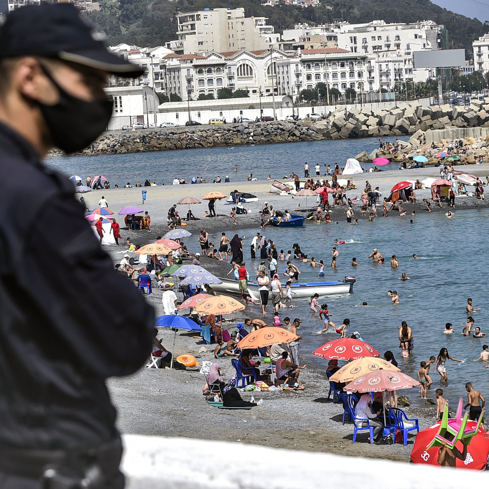 L'image montre un agent de sécurité en premier plan, portant un masque, surveillant une plage animée. En arrière-plan, des familles et des groupes de personnes profitent du temps ensoleillé, se baignant dans l'eau et se reposant sous des parasols colorés. On aperçoit également des bateaux au loin et des bâtiments sur la côte qui ajoutent à l'ambiance estivale de la scène.