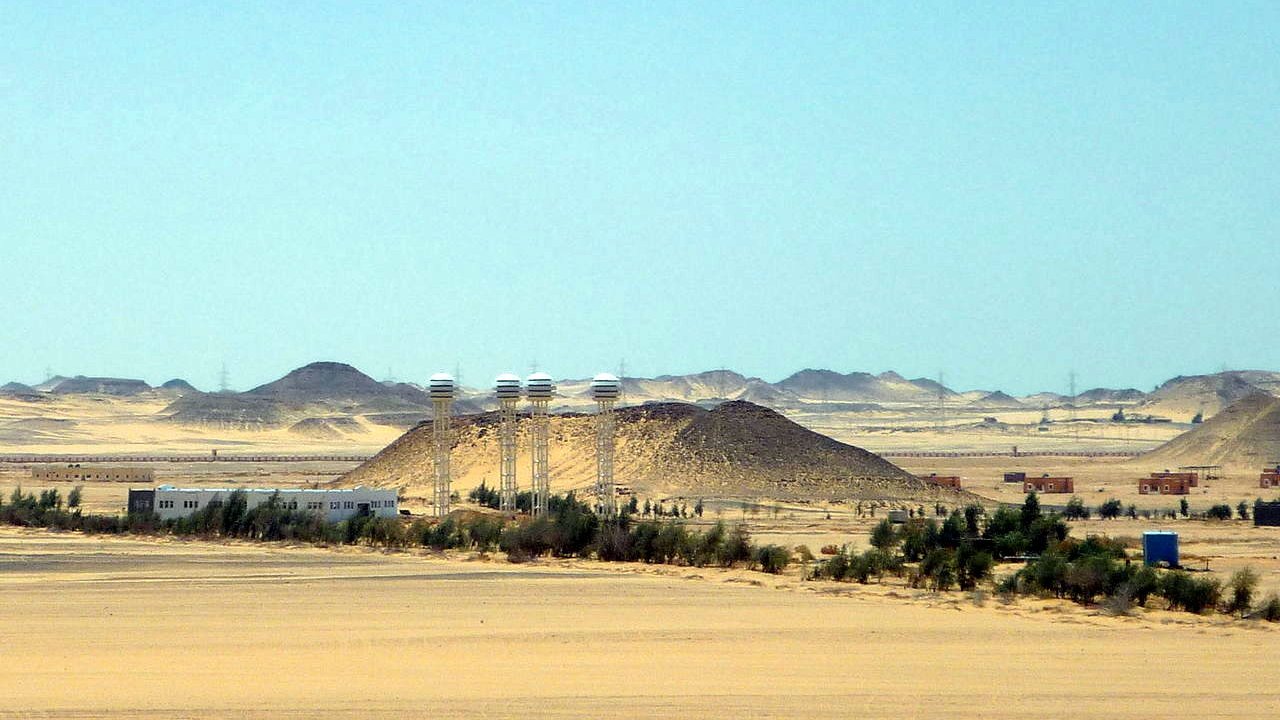 L'image montre un paysage désertique vaste et aride, avec des dunes de sable et des collines. Au centre, on peut distinguer quelques bâtiments qui semblent être des installations humaines, entourés de végétation telle que des arbres ou des buissons. À l'arrière-plan, des collines se découpent contre le ciel, tandis qu'une lumière vive souligne l'immensité du paysage. L'atmosphère semble calme et isolée, typique des régions désertiques.