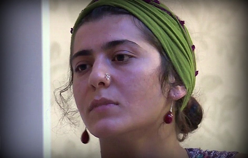 L'image montre une jeune femme avec un regard pensif. Elle porte un foulard vert sur la tête, orné de petites perles. Son visage est en partie encadré par des cheveux bouclés, et elle a un piercing au nez. Elle semble concentrée ou réfléchie, ce qui donne une ambiance contemplative à l'image.