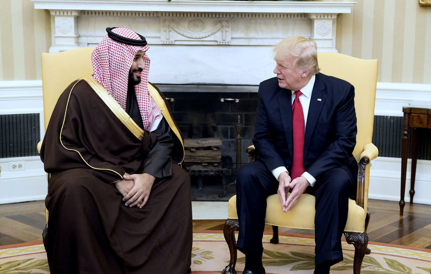 L'image montre deux hommes assis dans un salon élégant. Ils sont réunis dans un cadre formel, avec une cheminée en arrière-plan. L'un d'eux porte une tenue traditionnelle, comprenant un manteau et un headdress typique, tandis que l'autre est en costume. Ils semblent engagés dans une conversation amicale, affichant des expressions détendues. L'environnement est décoré de manière classique, avec des murs aux couleurs chaudes et quelques éléments de décoration.