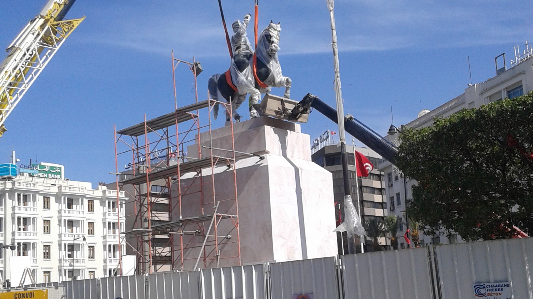 L'image montre un monument en cours d'installation. On peut voir une statue équestre accompagnée d'un cavalier, qui est en train d'être soulevée par une grue. Des échafaudages entourent la base de la statue, et un panneau de chantier est visible en arrière-plan. Le ciel est bleu, indiquant une journée ensoleillée. Des personnes marchent au premier plan, certaines portant des vêtements traditionnels.