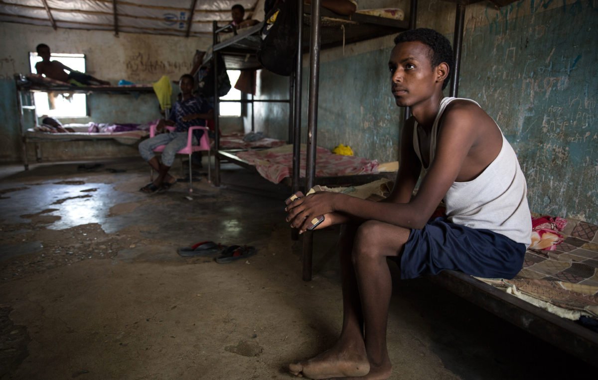 L'image montre un espace intérieur qui semble être une chambre ou un dortoir. On y voit plusieurs lits superposés et des personnes assises ou allongées. Un jeune homme est assis sur le bord d'un lit, regardant pensivement au loin. Il porte un débardeur et un short, et ses pieds sont nus. Les murs de la pièce sont nus et usés, avec des traces d'humidité, ce qui indique un environnement modeste et probablement surpeuplé. La lumière naturelle pénètre par une fenêtre, ajoutant une certaine ambiance à la scène.