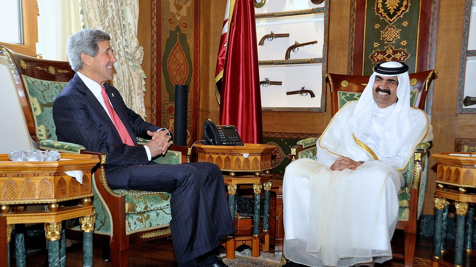 L'image montre deux hommes assis dans un bureau élégant, décoré avec des motifs riches et des couleurs vives. L'un des hommes porte un costume formel et sourit, tandis que l'autre est vêtu d'une tenue traditionnelle blanche, avec un chapeau noir. Ils semblent être en train de discuter dans une atmosphère amicale. En arrière-plan, on peut voir des objets décoratifs et des armes anciennes accrochées au mur, ajoutant à l'ambiance opulente de la pièce.