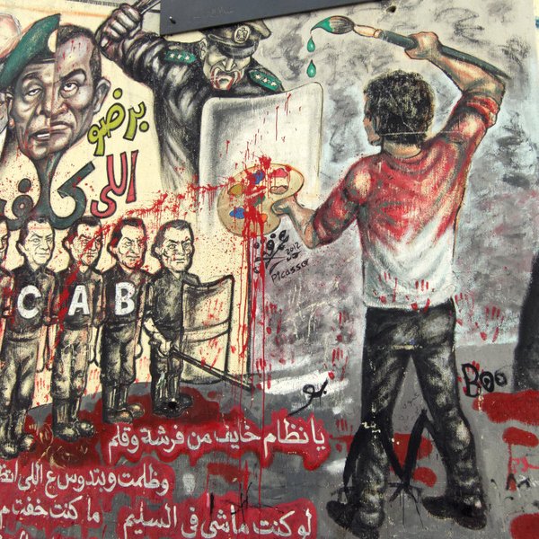 L'image montre un mural coloré, rempli de graffitis et d'illustrations. On y voit une scène dynamique où une personne, de dos, est en train de peindre avec un pinceau, probablement en train d'exprimer un message de frustration ou de résistance. Autour de cette figure, il y a diverses représentations de policiers et de figures autoritaires, certains avec des visages mécontents, indiquant une atmosphère de contestation. Le mur est éclaboussé de peinture rouge, ce qui pourrait symboliser la violence ou le sang, renforçant le message de lutte contre la répression. Les lettres "ACAB" sont visibles, ce qui fait référence à une phrase souvent associée à la critique des forces de police. L'ensemble de la composition semble porter un message fort sur l'opposition à l'autorité.