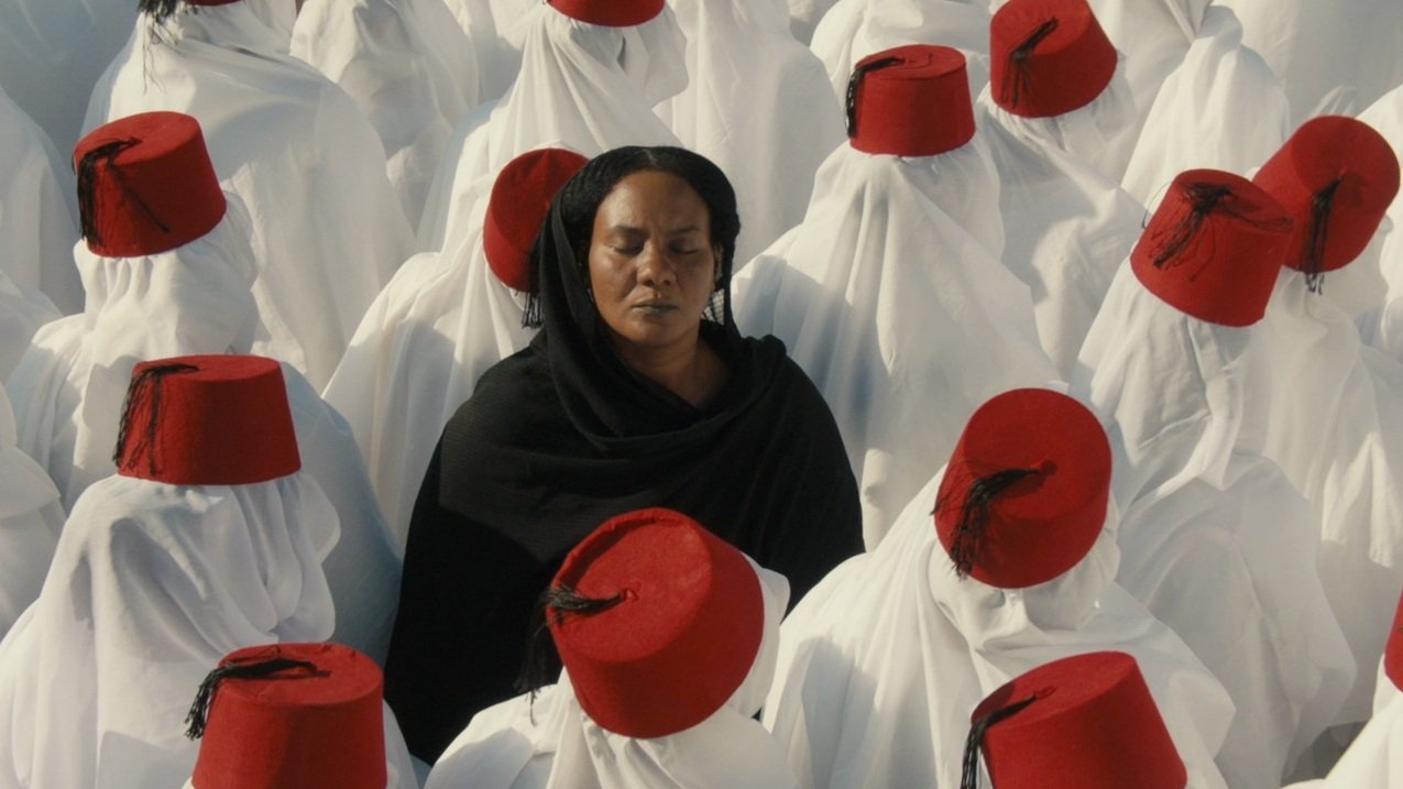 L'image montre un groupe de personnes vêtues de longues robes blanches, portant des fez rouges. Au centre, une femme en noir se tient les yeux fermés, contrastant avec l'uniformité des autres. Cette composition crée une atmosphère de méditation ou de réflexion, avec un fort accent sur la spiritualité et la communauté. Les couleurs et les vêtements évoquent une tradition culturelle riche.