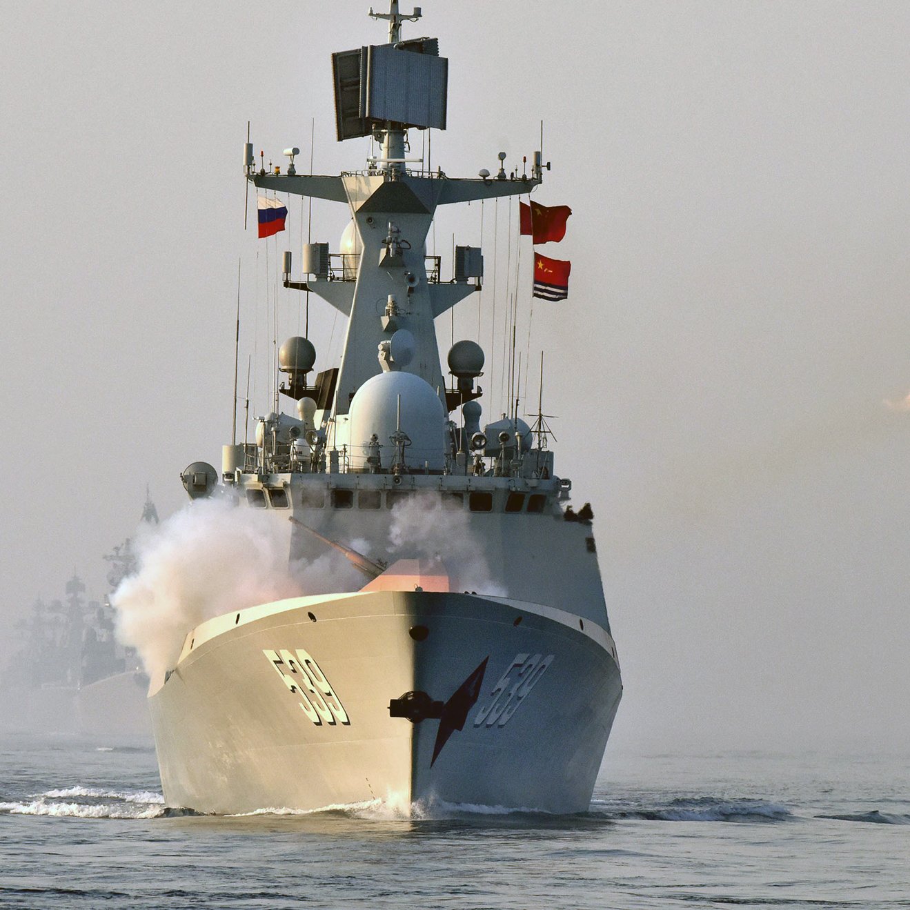 L'image montre un navire de guerre en mer, en train de tirer un missile. On peut voir de la fumée s'échapper du projetile, tandis que le bateau est orné de plusieurs drapeaux. L'atmosphère semble brumeuse, ce qui ajoute une certaine dramatisation à la scène. En arrière-plan, d'autres navires sont indistinctement visibles.