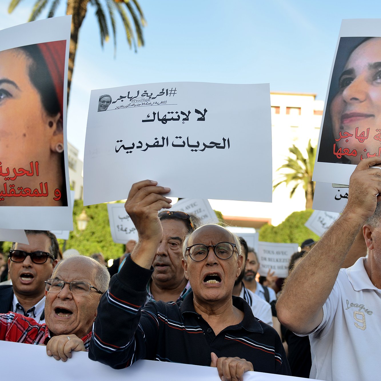 L'image montre une manifestation avec un groupe de personnes tenant des pancartes. Les manifestants semblent exprimer leur désaccord et revendiquer des droits, notamment en ce qui concerne les libertés individuelles. Certains affichent des expressions de colère ou de détermination, tandis que d'autres portent des signes clairs de solidarité. En arrière-plan, on peut voir des palmiers et un bâtiment, suggérant un cadre urbain. L'atmosphère est engagée et militante, reflétant une lutte pour des causes sociales ou politiques.