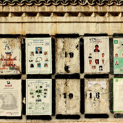 L'image montre un mur sur lequel sont affichés plusieurs documents et affiches. Ces derniers semblent être en lien avec des élections, avec des plaques présentant des photos et des noms de candidats. Certaines affiches sont abîmées et décolorées, révélant un certain vieillissement et une usure du temps. Le mur lui-même présente une texture rugueuse et des motifs décoratifs en haut. Les divers éléments placardés créent une composition visuelle chargée et informative.