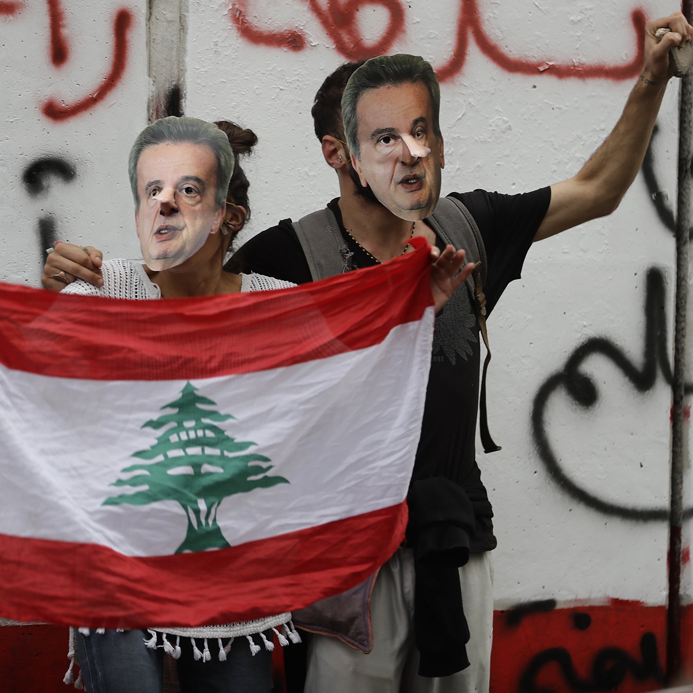 L'image montre deux personnes tenant un drapeau libanais tout en portant des masques représentant un homme. Ils se tiennent devant un mur avec des inscriptions en arabe, probablement dans un contexte de manifestation ou de protestation. L'ambiance semble chargée d'émotion et de revendications politiques.