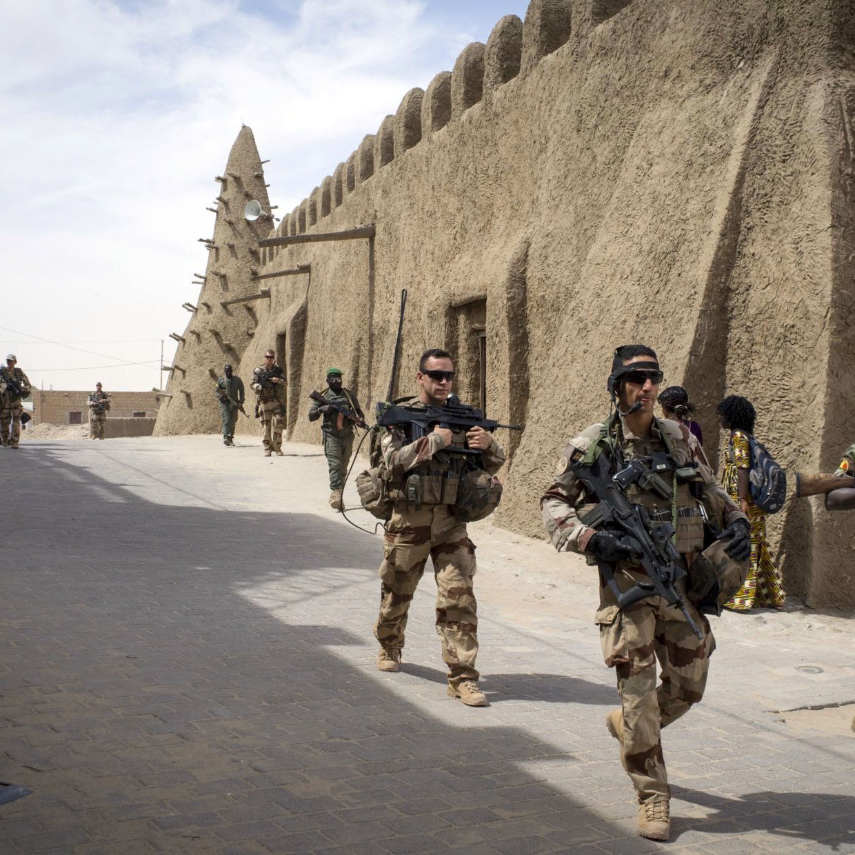 The image depicts a group of soldiers walking through a narrow street in a fortified area. They are dressed in military gear and armed with rifles. The architecture features traditional, mud-brick structures, which create a historical and cultural backdrop. The scene conveys a sense of military presence and activity, with more soldiers seen in the background, indicating a coordinated operation or patrol. The atmosphere appears to be tense and vigilant, typical of a military environment.