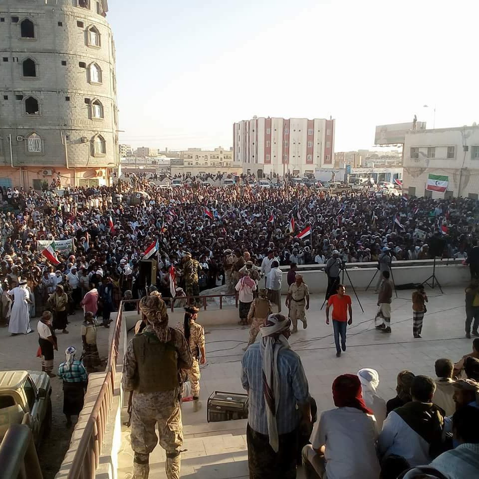 در این تصویر، یک تجمع یا راهپیمایی بزرگ در فضای باز مشاهده می‌شود. مردم زیادی در مقابل یک ساختمان قرار دارند و برخی از آن‌ها پرچم‌هایی به دست دارند. در پس‌زمینه، ساختمانی بزرگ و نیمه‌ساز دیده می‌شود، و ساختمان‌های دیگری نیز در دوردست‌ها وجود دارد. نور خورشید به صورت عمودی به این منطقه می‌تابد و جو عمومی نشان‌دهنده شور و هیجان مردم است. گروهی از افراد در جلو و عده‌ای دیگر در پشت آن‌ها نشسته‌اند یا ایستاده‌اند.