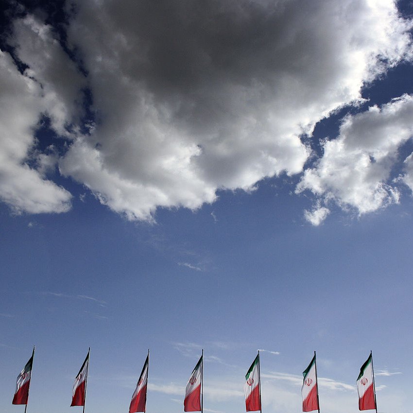 The image features a clear blue sky with fluffy white clouds. In the foreground, several flags, likely representing Mexico, are lined up. The flags prominently display the colors of the Mexican flag, which includes green, white, and red, along with the national emblem. The overall scene conveys a sense of patriotism against a serene backdrop.