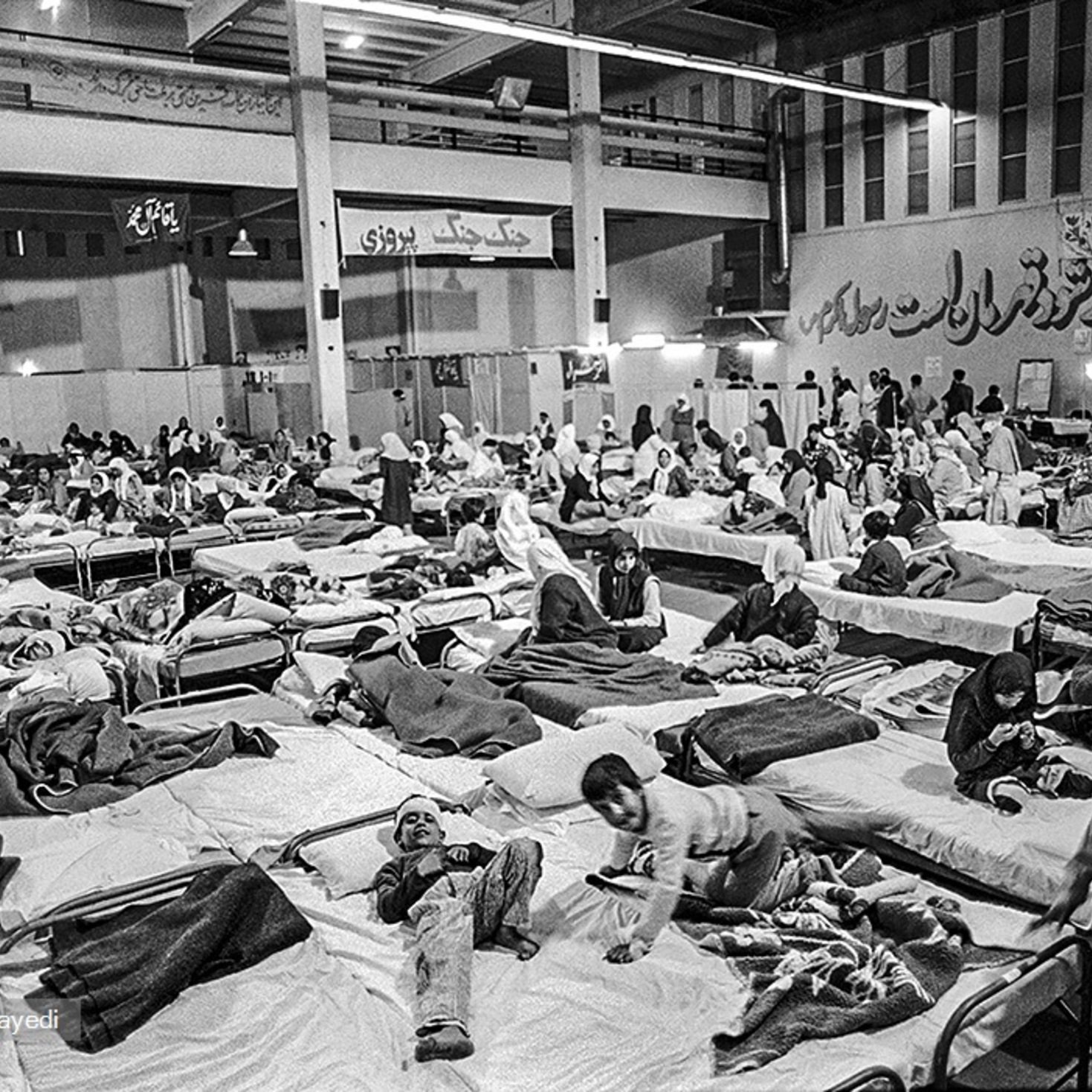 L'image montre un grand espace intérieur aménagé en salle de soins, avec de nombreux lits occupés par des personnes. On peut voir des patients allongés sur des matelas, certains semblant recevoir des soins médicaux. L'atmosphère est chargée et il y a un sentiment d'urgence, avec une présence significative de personnel soignant et de familles. Les murs portent des inscriptions en persan, ajoutant à l'authenticité du lieu.