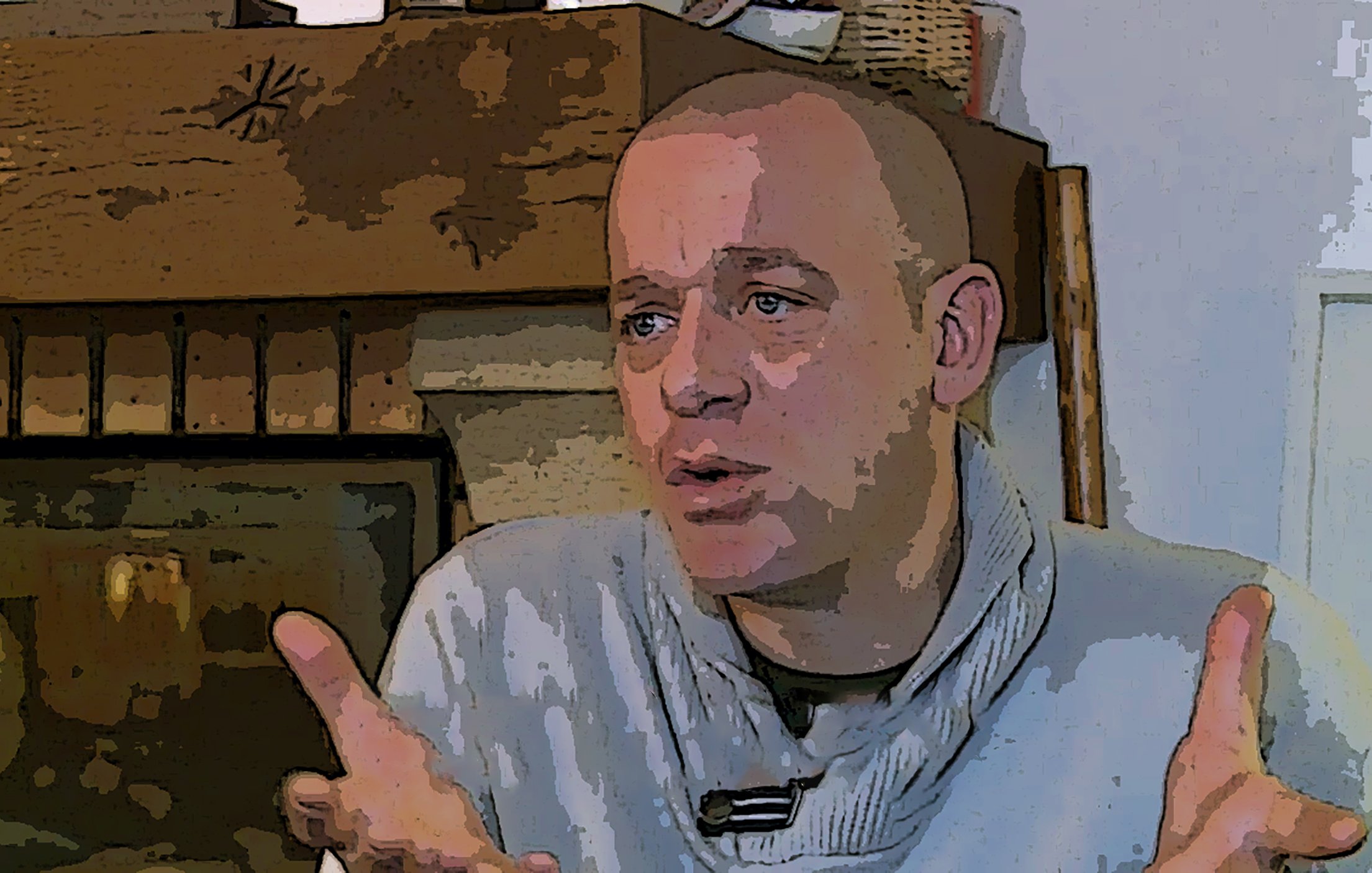 L'image montre un homme avec une coupe de cheveux courte et un pull en tricot. Il est assis près d'une cheminée en pierre. Son expression faciale semble être sérieuse, et il gesticule avec ses mains, ce qui suggère qu'il est en train d'expliquer ou de discuter d'un sujet important. L'arrière-plan présente des éléments décoratifs, ce qui ajoute une ambiance chaleureuse à la scène.