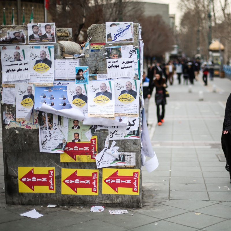 تظهر الصورة مشهداً في منطقة حضرية، حيث تُظهر مجموعة من الملصقات الإعلانية المعلقة على جدار، تتضمن صوراً وعبارات مكتوبة. في المقدمة، توجد امرأة ترتدي حجاباً أسود، تسير في اتجاه معين. الطريق مزدحم بعض الشيء، مع وجود أشخاص آخرين في الخلفية ووسائل النقل. يبدو أن الجو يميل إلى البرودة، ويمكن رؤية الأشجار والبنية التحتية الحضرية.